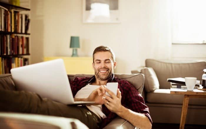 A man relaxing on a couch, smiling while using his smartphone with a laptop on his lap. Blackout shades frame the cozy living room, where a bookshelf, lamp, and coffee table add to the inviting atmosphere.
