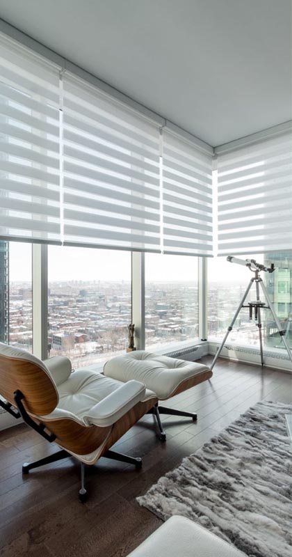 A modern lounge with floor-to-ceiling windows, featuring a white leather chair with matching ottoman, a telescope, striped blinds, and city views. Blackout shades offer privacy and style while a soft gray rug covers part of the wooden floor.