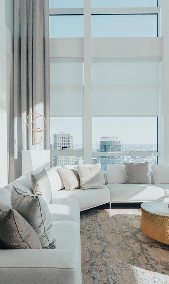 Modern living room with large windows featuring sleek exterior window shades, a white sectional sofa with gray and beige pillows, a patterned rug, and a gold-toned coffee table. City buildings are visible outside in the bright natural light.