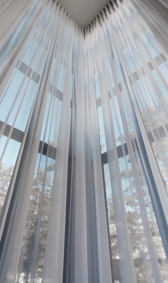 Sheer white curtains serve as elegant windows treatments, hanging from ceiling to floor in the corner of a bright room, with sunlight streaming through large windows and trees faintly visible outside.