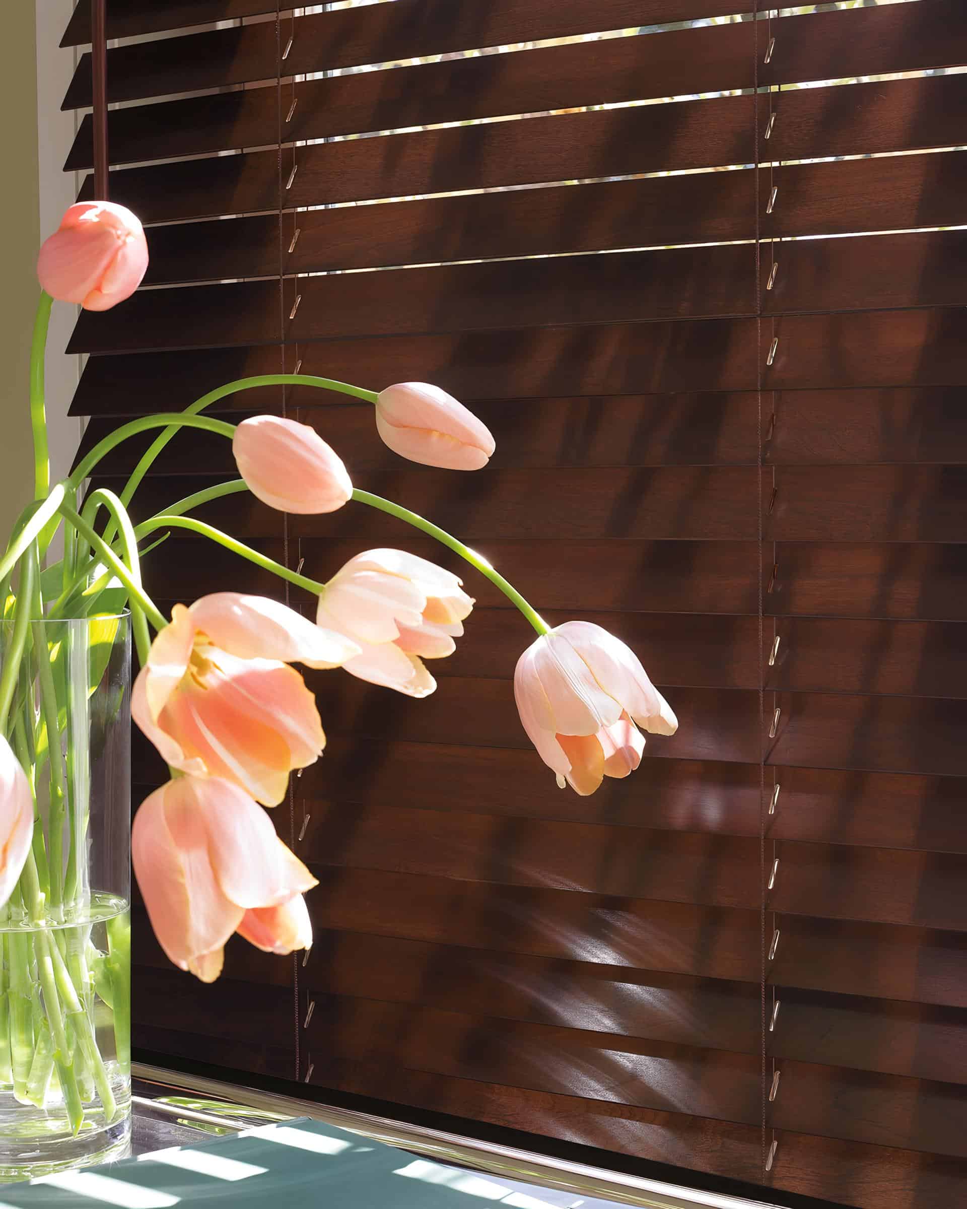 A glass vase with drooping pink tulips sits in front of a window featuring dark wooden blinds, sunlight casting soft shadows across the flowers and expertly installed windows coverings.