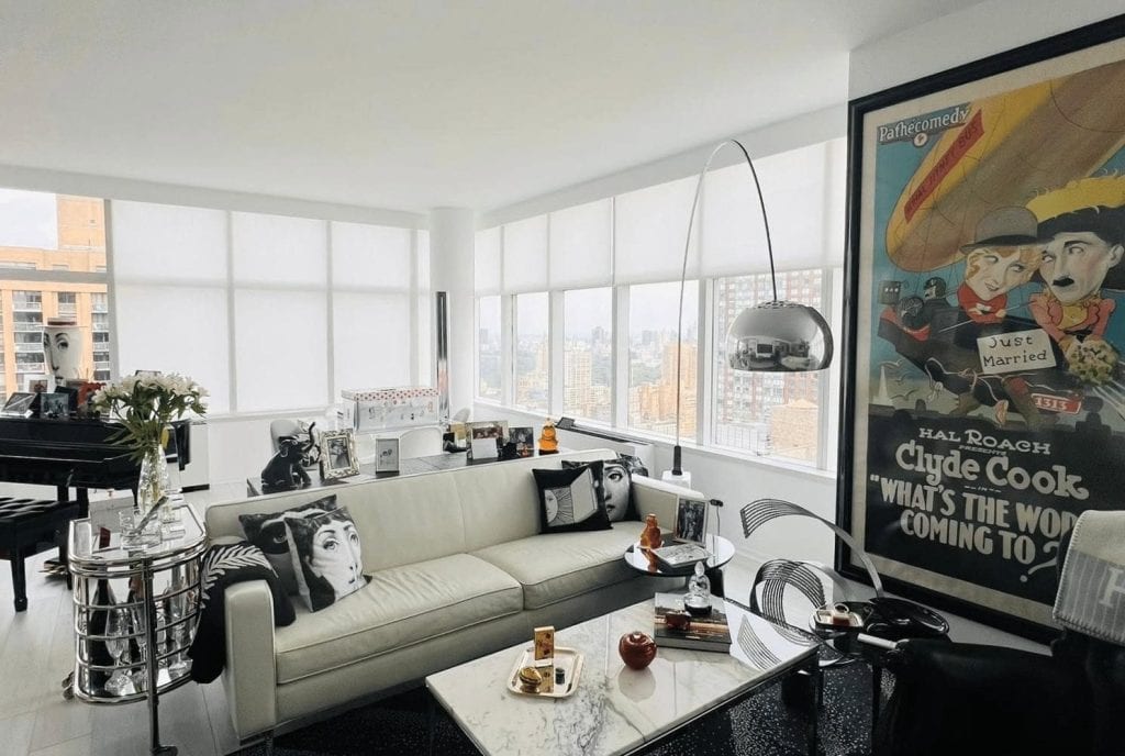 A bright, modern living room with large windows, a white sofa with classic film-themed pillows, a marble coffee table, a piano, and a vintage movie poster on the wall. City buildings are visible outside.