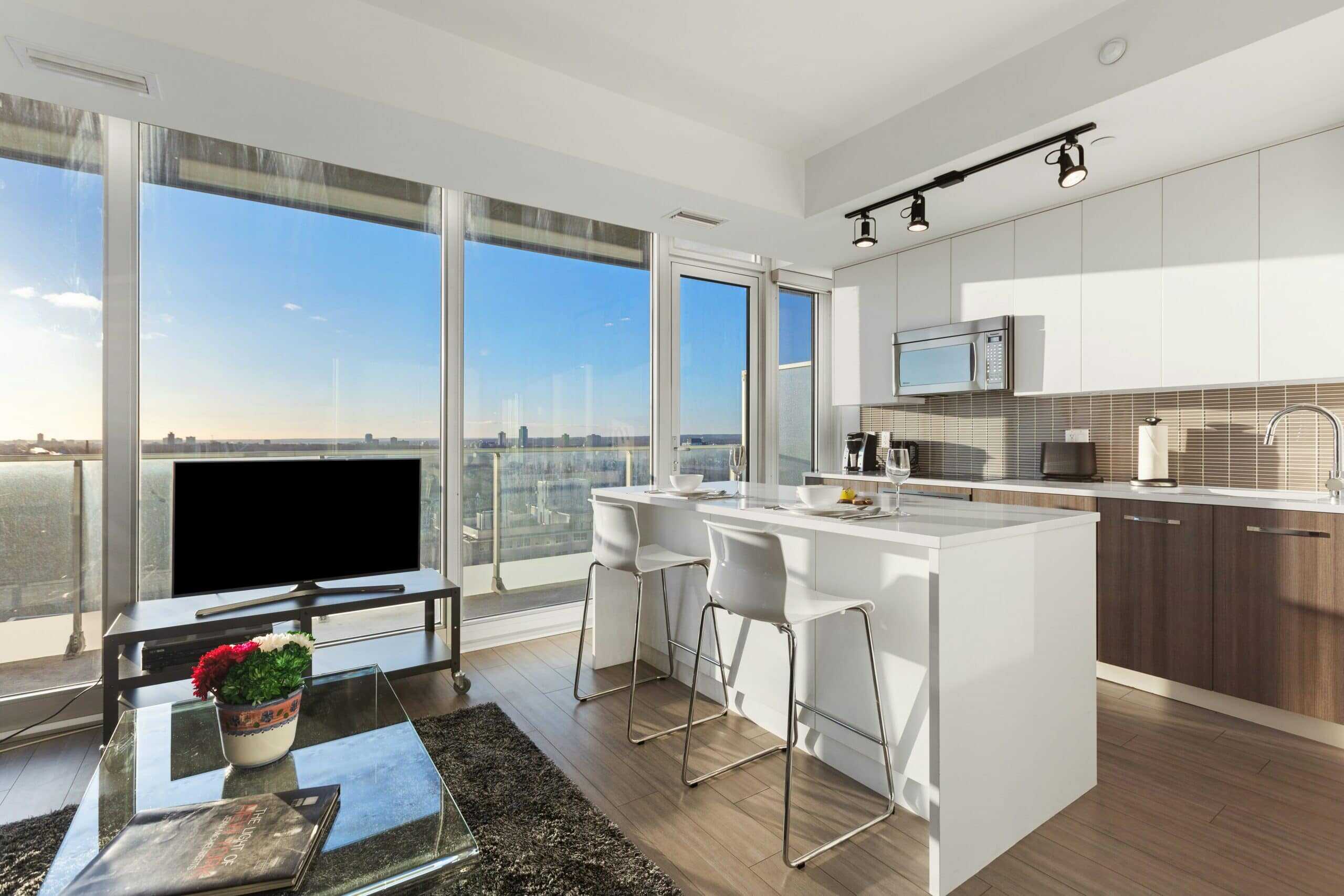 Modern apartment with floor-to-ceiling windows featuring sleek window coverings, a kitchen with white cabinets and island, a TV on a stand, glass coffee table with a plant, and stunning city views outside.