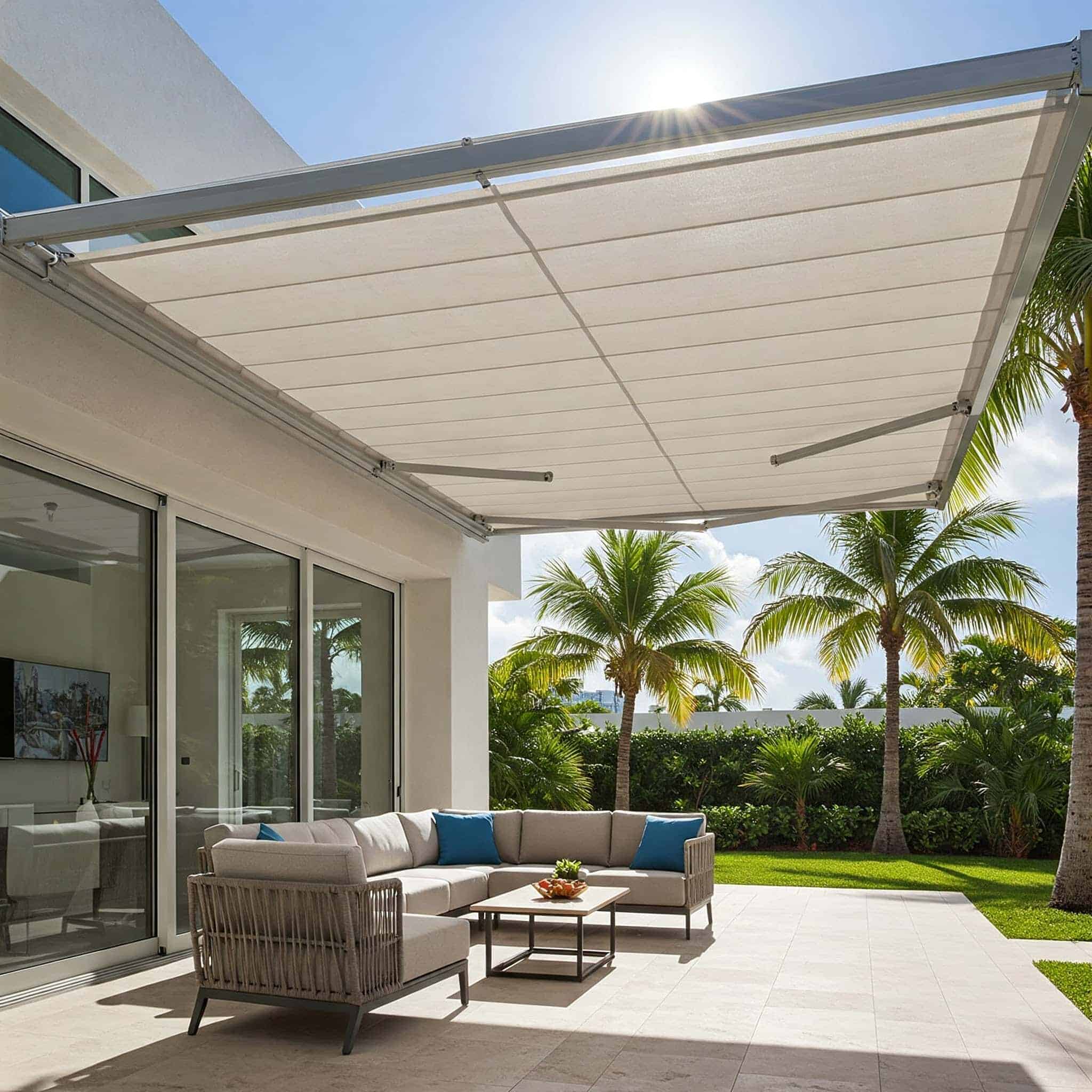 A modern outdoor patio with a large retractable awning, beige sectional sofa, coffee table, and palm trees in the background under a sunny blue sky, features sleek exterior window shades for added comfort and style.