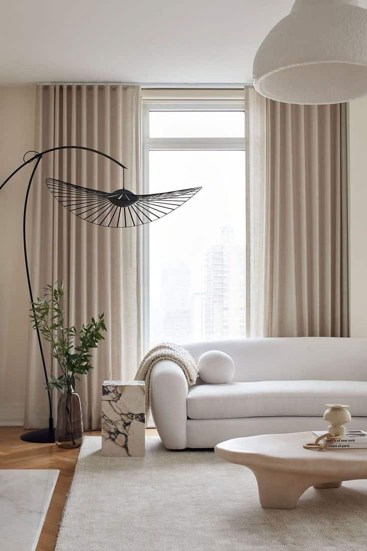 A modern living room with a white curved sofa, a large floor lamp with a black wire shade, beige curtains by an expert blinds installer, a marble side table, low coffee table, and a potted plant by the window.