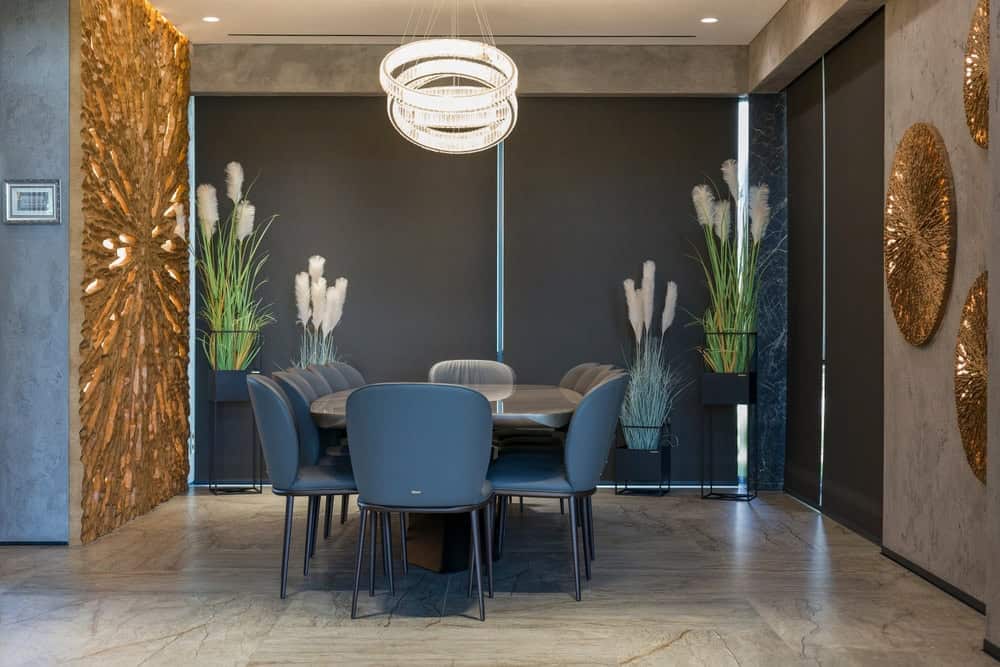 Modern dining room with a large dark table, eight gray chairs, circular chandeliers above, black window shades, decorative plants, and gold abstract wall art on textured gray walls and floor.