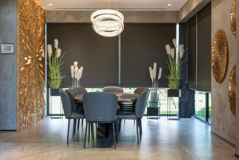 Modern dining room with a black table, gray chairs, large black window blinds, potted tall grasses, gold textured wall art, and a circular chandelier hanging above the table.