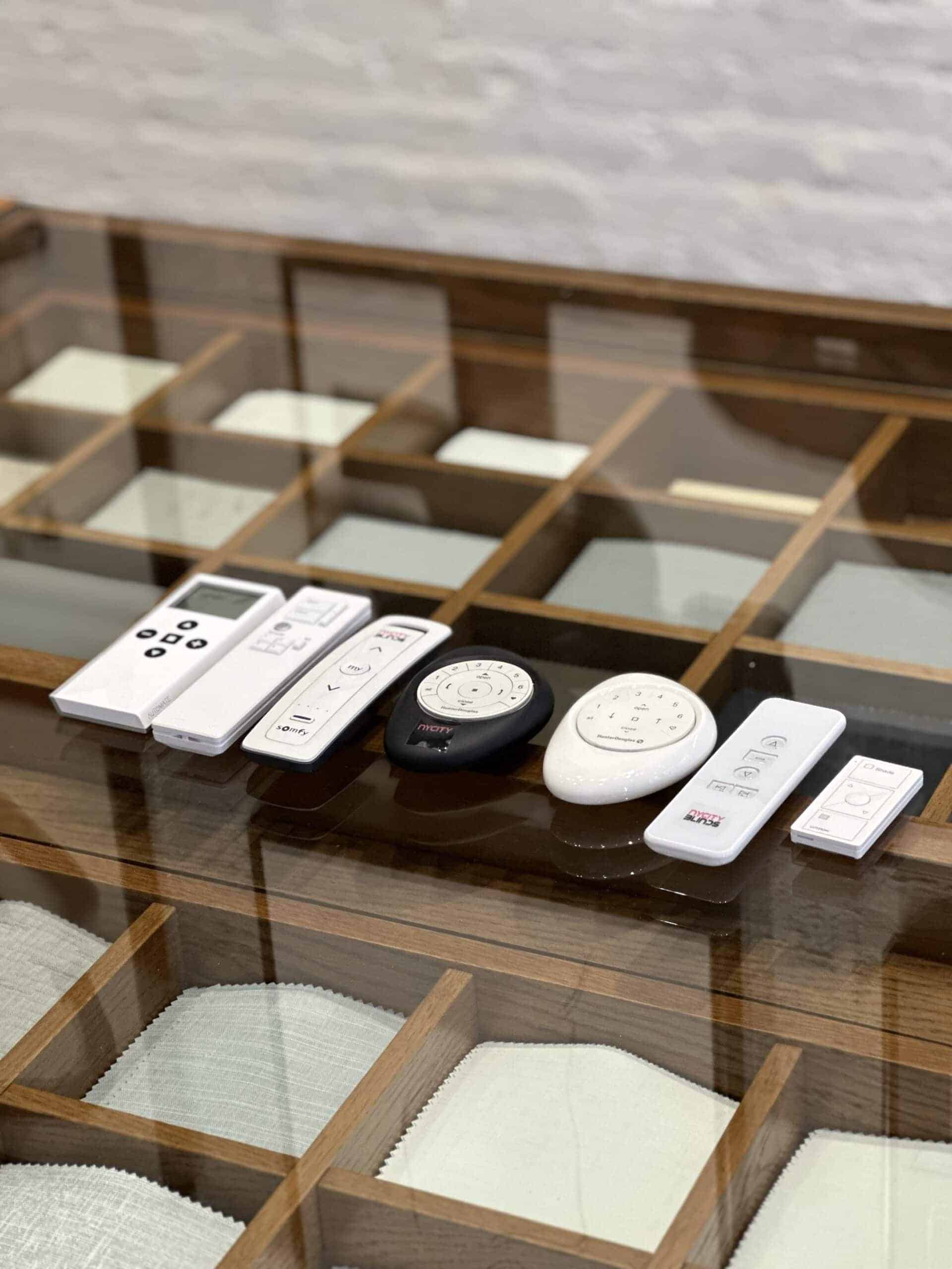 A variety of remote controls, mostly white and one black, are neatly arranged in a row on a glass display case with wooden dividers below.