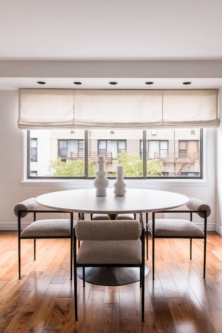 A modern dining area with a round white table, four upholstered chairs, and three white vases. Large windows with cream Roman shades provide natural light, overlooking an urban building and trees.