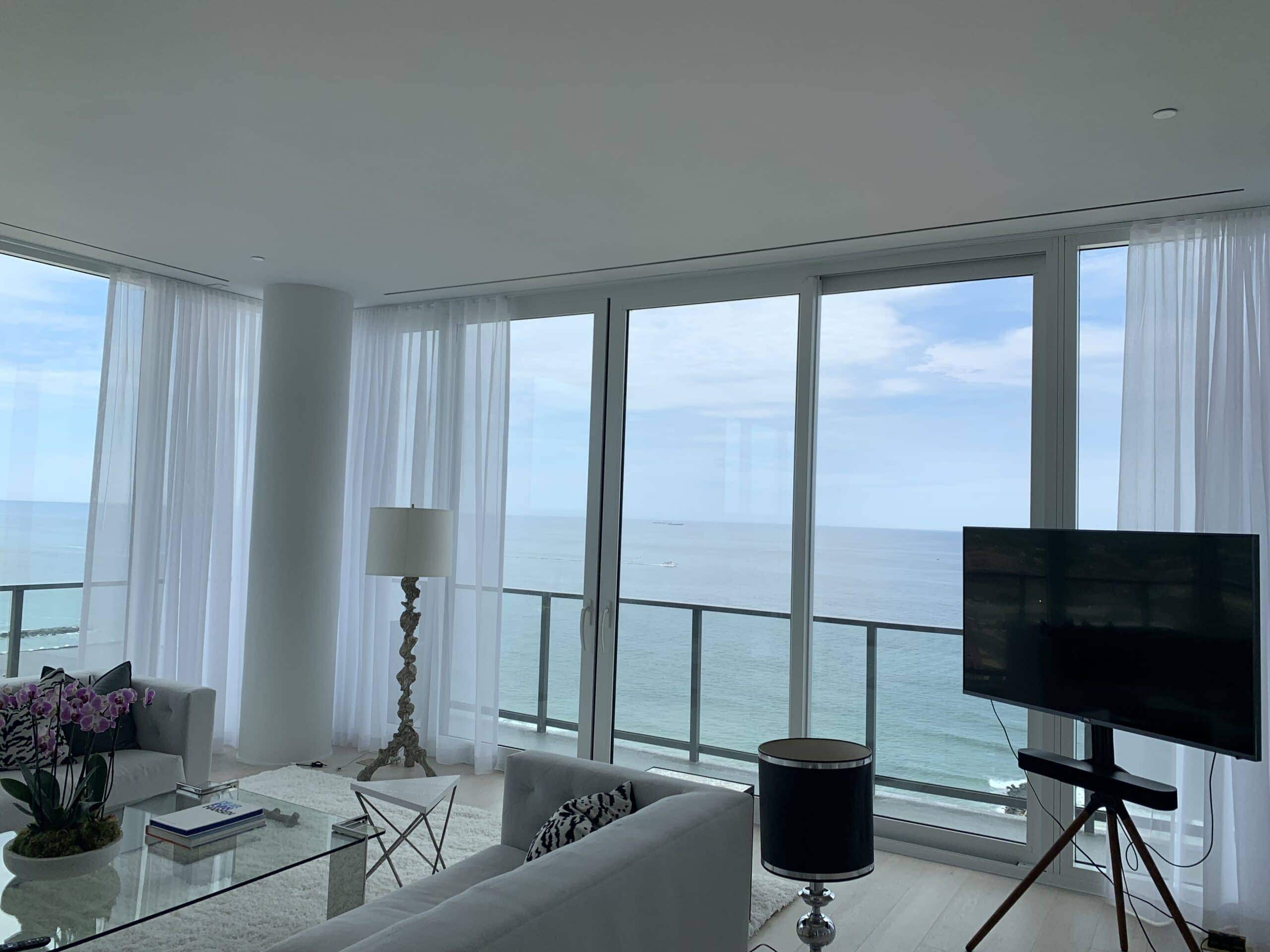 A modern living room with large floor-to-ceiling windows offers a panoramic ocean view. White furniture, a glass coffee table with flowers, a TV on a stand, and sheer curtains complete the airy, bright space.