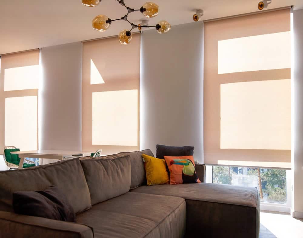 A modern living room with a gray sectional sofa, colorful cushions, and large windows covered by beige roller blinds. Warm sunlight filters through the blinds, creating a cozy and inviting atmosphere.