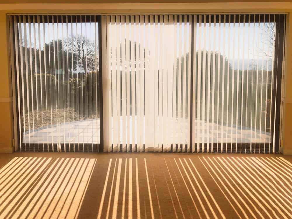 Vertical blinds cover large glass doors, with sunlight streaming through and casting striped shadows across a beige carpeted floor. A patio and greenery are visible outside in bright, sunny weather.