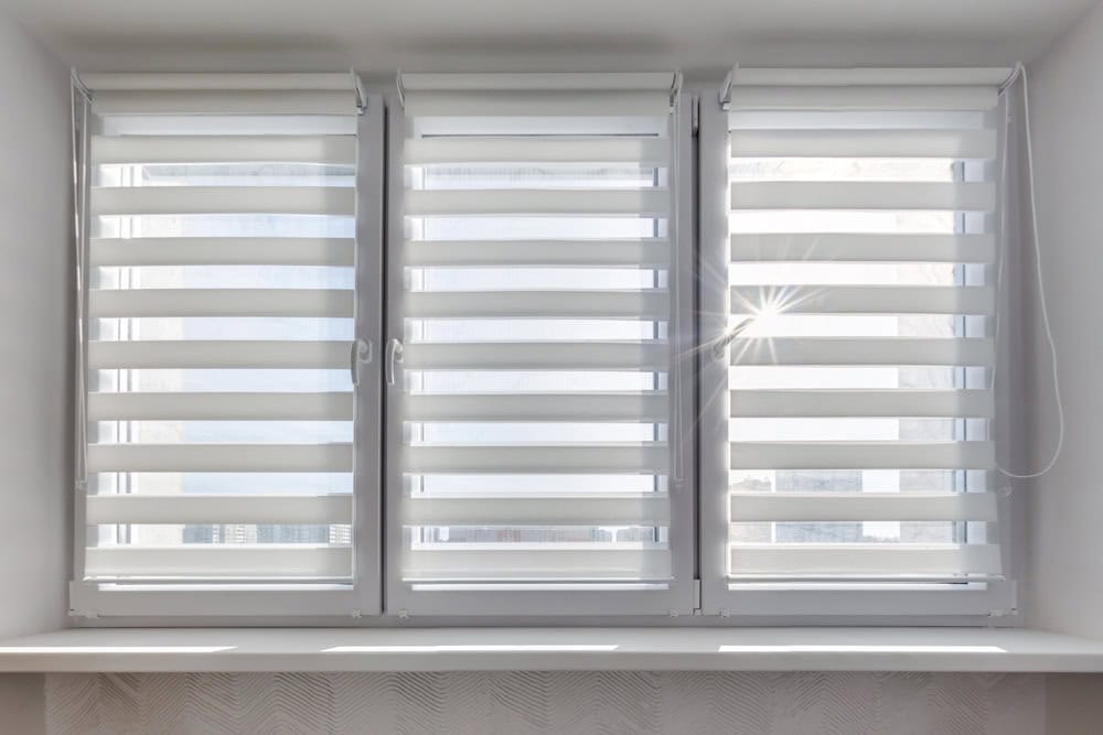 Three large windows with white zebra blinds partially open, allowing sunlight to filter through, creating striped light patterns. A bright sunbeam is visible through the middle window.