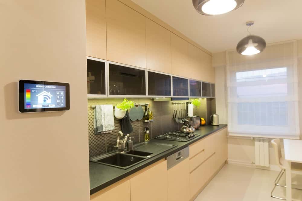 Modern kitchen with light wood cabinets, black countertops, and a smart home control panel on the wall. Kitchen appliances and utensils are neatly arranged, with pendant lights and a large window providing natural light.