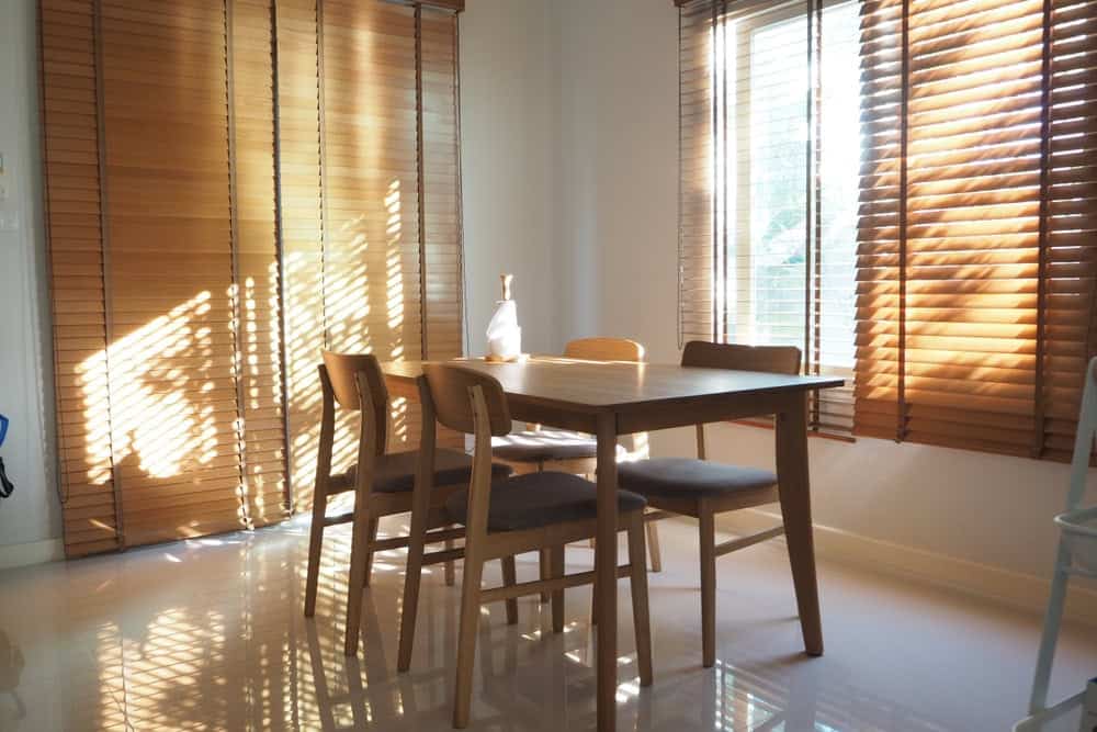 A sunlit dining area with a wooden table and four chairs, placed near large windows with wooden blinds casting shadows across the polished floor. A decorative bottle sits on the table.