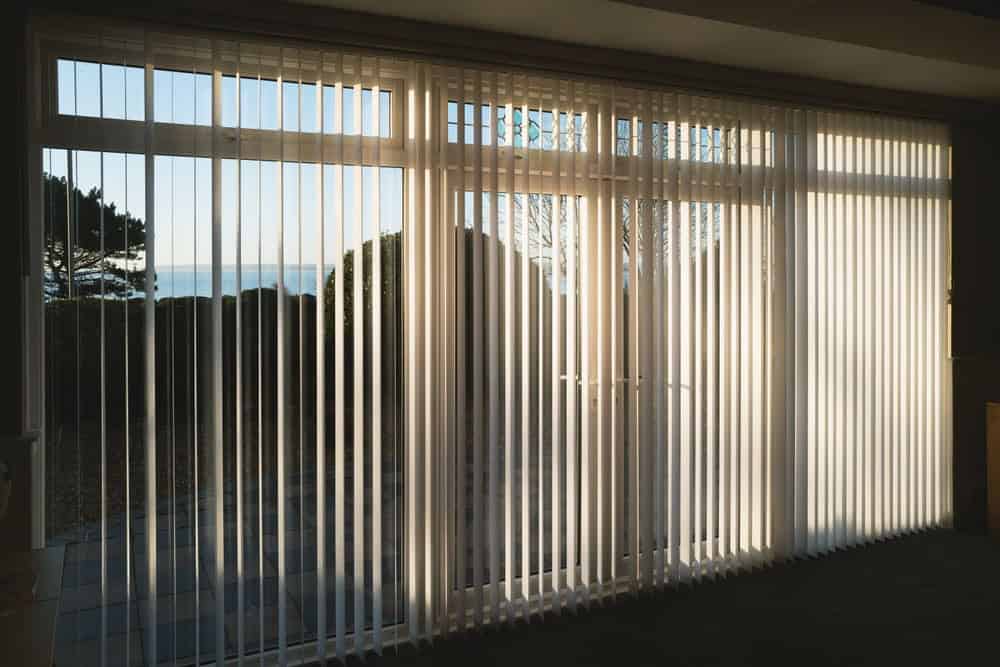 Vertical blinds cover large glass doors, letting in sunlight. Outside, there’s a patio, bushes, and a tree, with a view of the sea and sky in the background. Sunlight casts striped shadows on the floor.