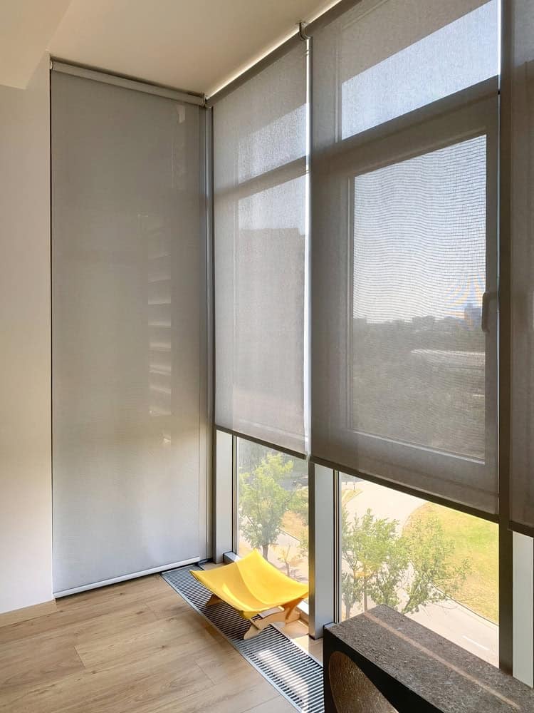 A bright room with large floor-to-ceiling windows covered by translucent blinds. A modern yellow lounge chair sits near the window, and trees and grass are visible outside. Light wood flooring completes the space.