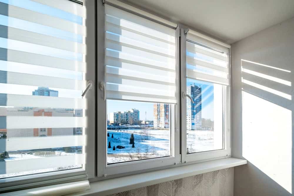 Three large windows with white zebra blinds partially open, letting in sunlight. Outside, there are modern apartment buildings and a snowy landscape with trees and blue sky. Light and shadow patterns fall on the wall and windowsill.
