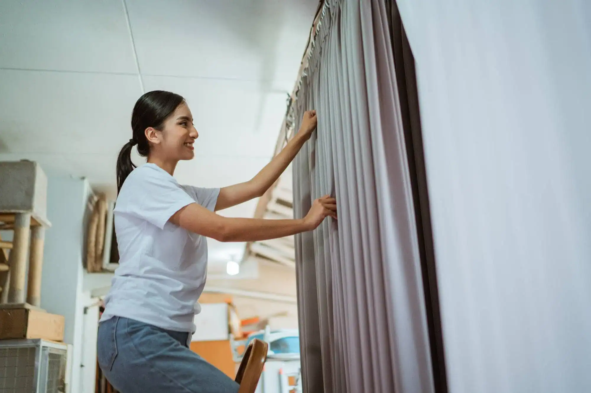 A woman standing on a stool is smiling while closing grey curtains, showcasing stylish windows treatments Miami-Dade County residents love. The well-lit FL room features shelves and various items in the background. She wears a white t-shirt and blue pants.