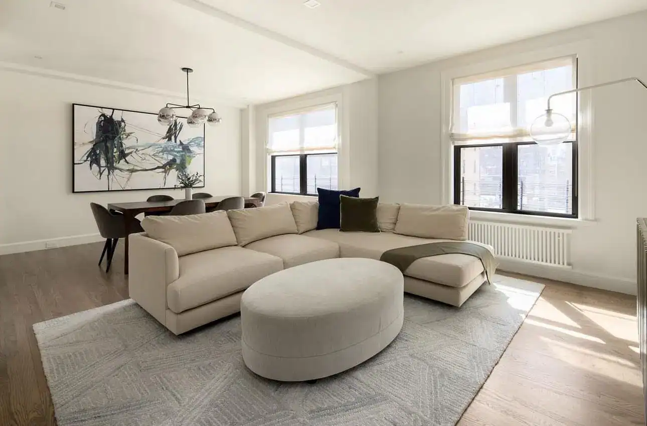 A modern living room with a beige sectional sofa, oval ottoman, large abstract painting, dining table with dark chairs, and large windows letting in natural light on wooden floors with a light area rug.