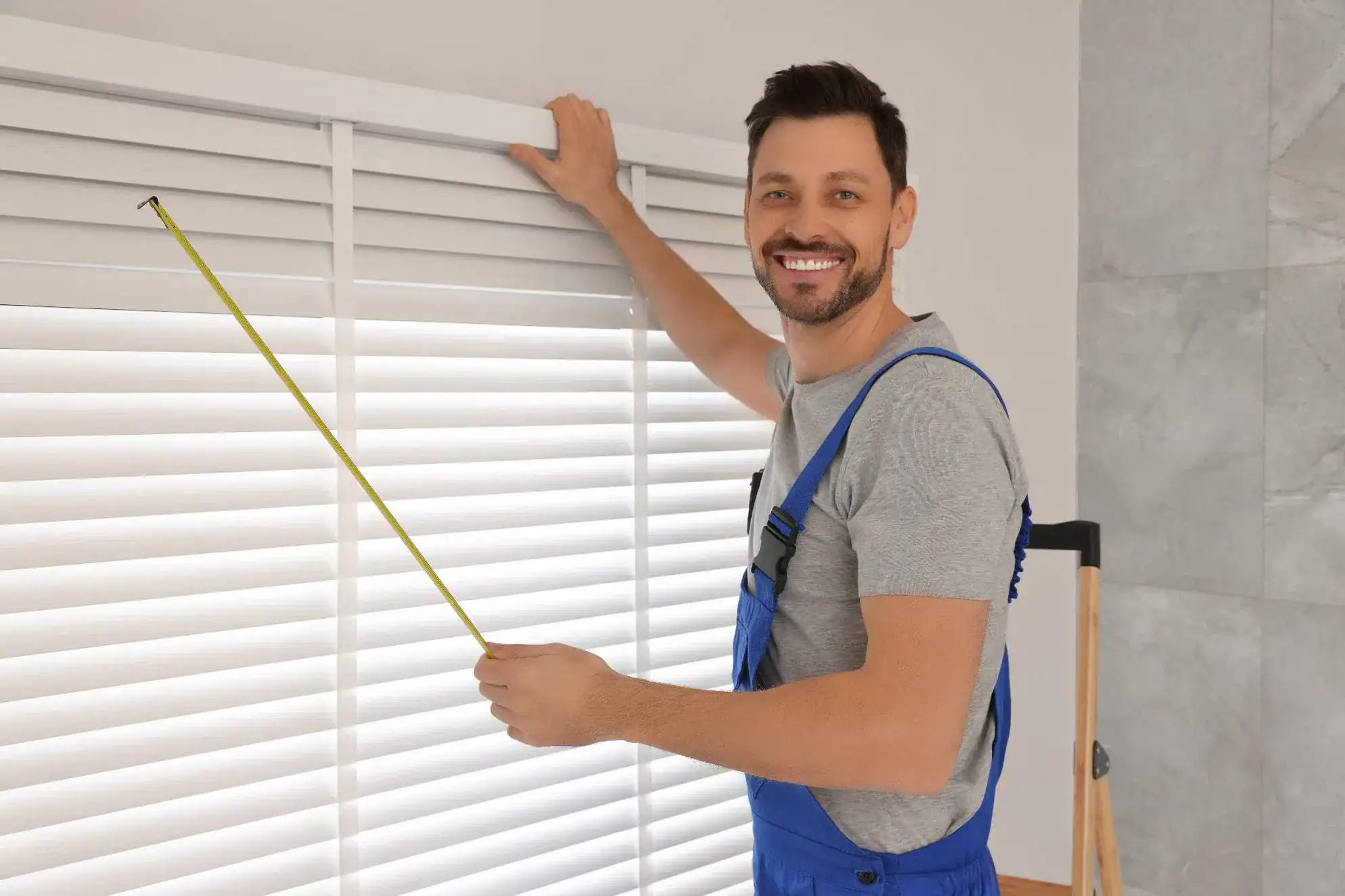 A smiling man in blue overalls measures a window with a tape measure while adjusting white horizontal blinds, showcasing expert windows treatments in Miami-Dade County, FL, in a modern, bright room.