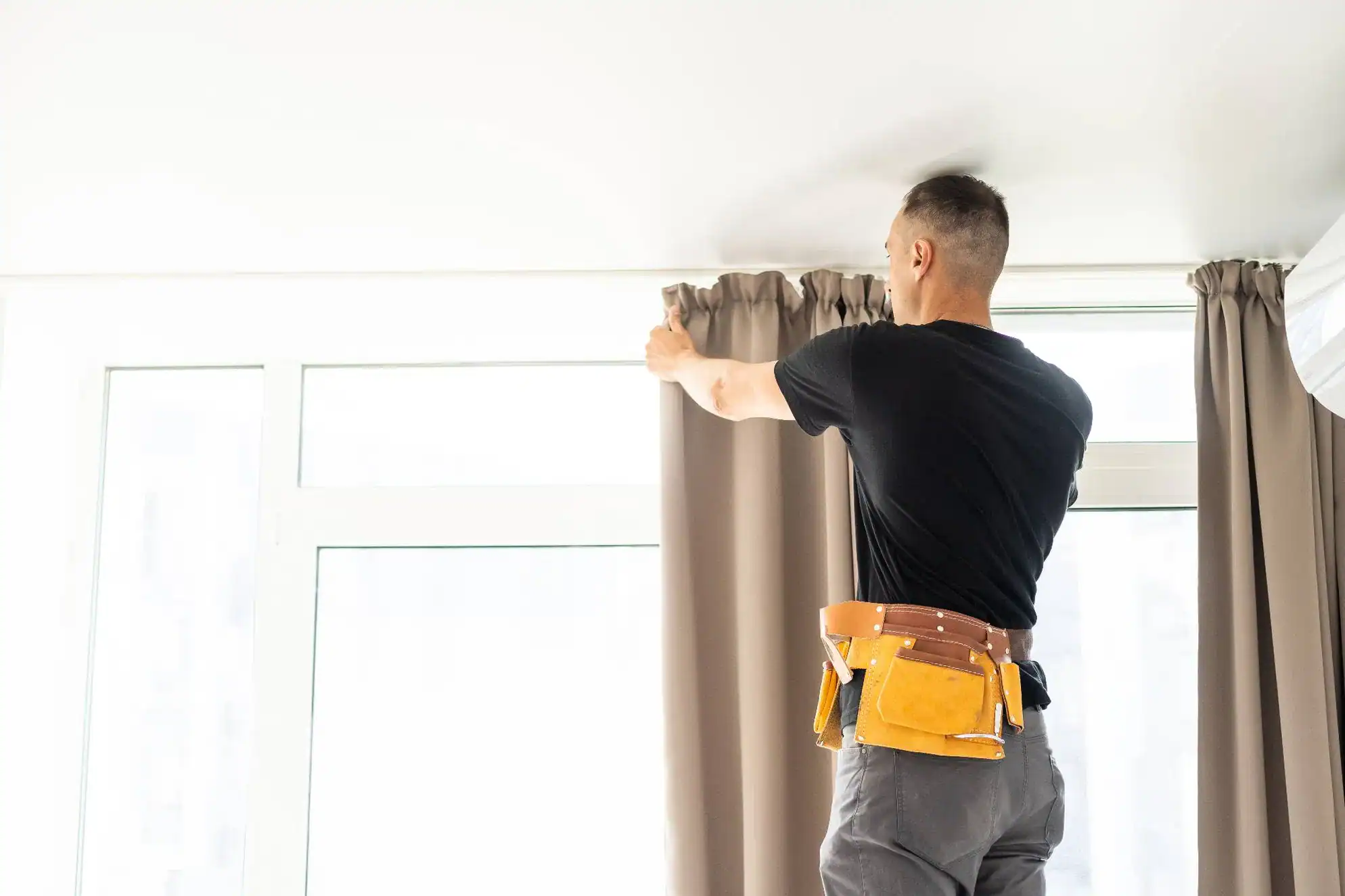 A person wearing a tool belt adjusts beige curtains—a popular choice for windows treatments Miami-Dade County—in a bright FL room, standing on the floor with their back to the camera.