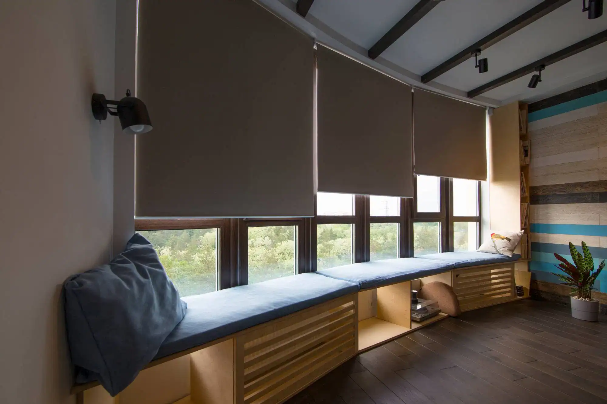 A modern room with large windows featuring sleek roller blinds, a cushioned window seat with pillows, wooden storage underneath, and a plant in the corner. Stylish wood panels and dark flooring highlight expert windows treatments Miami-Dade County, FL.