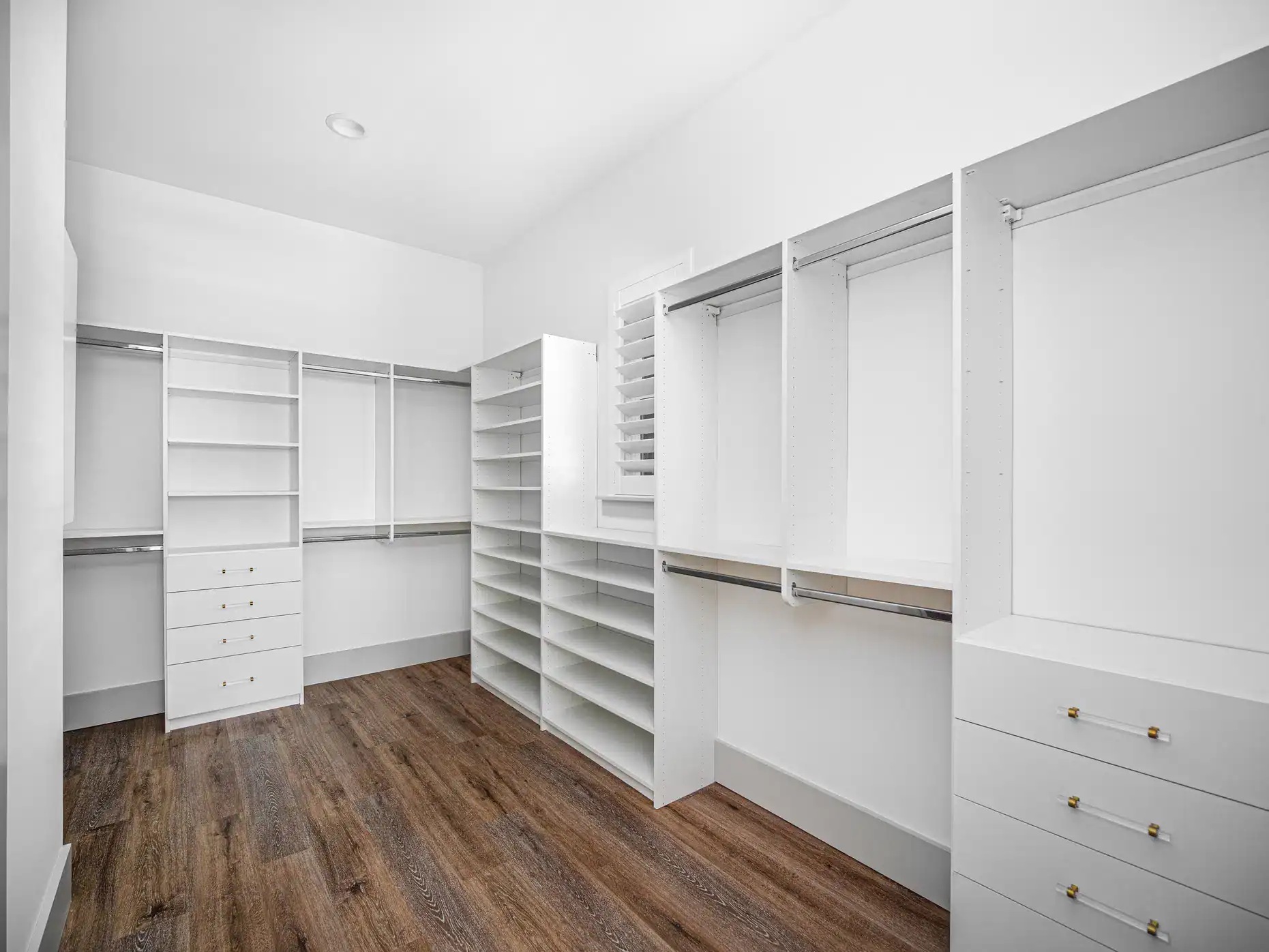 Spacious, modern walk-in closet with white built-in shelves, drawers, and hanging rods, set against a white wall with a wooden floor and stylish FL window treatments in the small window with blinds.
