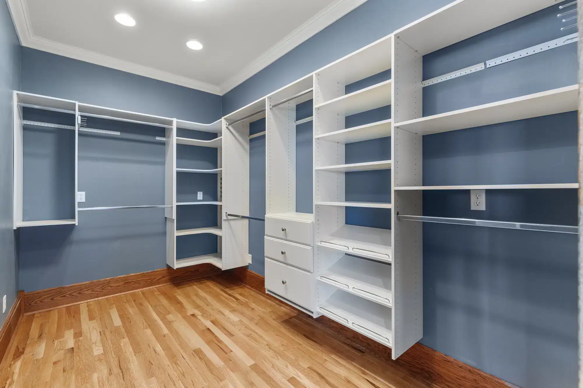 Spacious walk-in closet with blue walls, wooden floor, white built-in shelves, drawers, and multiple hanging rods, illuminated by ceiling lights—perfect for adding custom windows treatments in Miami-Dade County, FL.