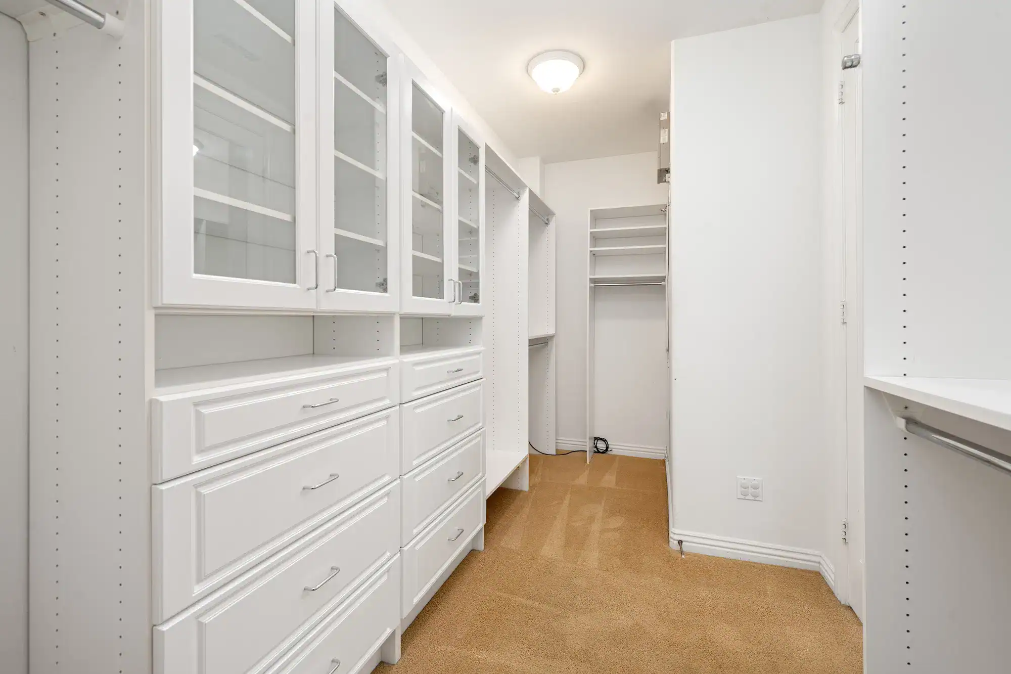 A spacious walk-in closet with white built-in shelves, glass-front cabinets, and drawers on one side. The tan carpeted floor and bright ceiling light create a welcoming space, perfect for showcasing elegant windows treatments Miami-Dade County, FL style.