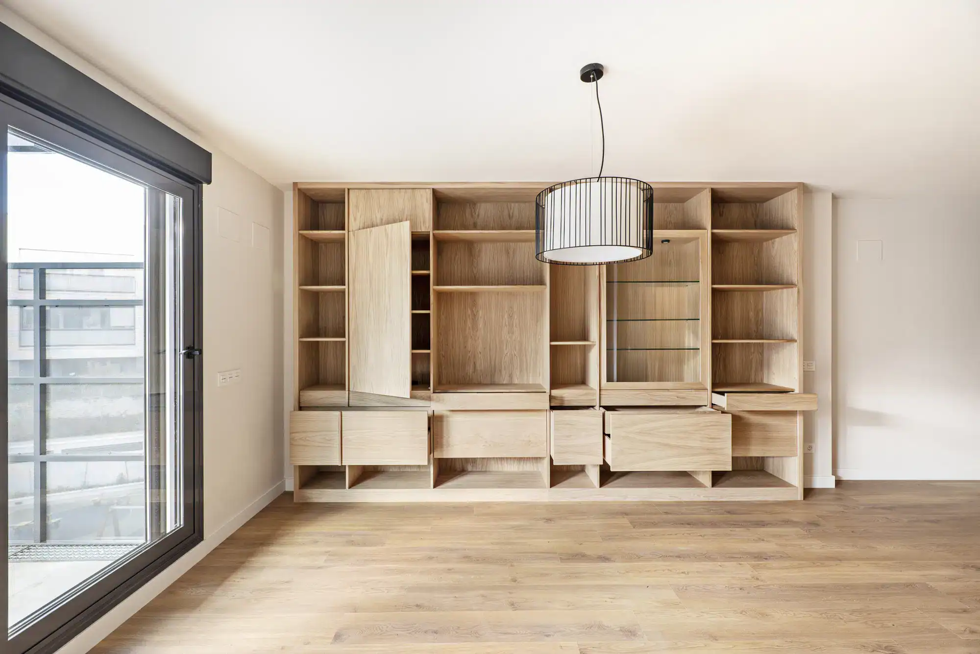 A modern, empty room with wood flooring, a large window on the left featuring stylish windows treatments Miami-Dade County, and a built-in wooden shelving unit. A black pendant light hangs from the ceiling. Ideal FL living with elegant details.