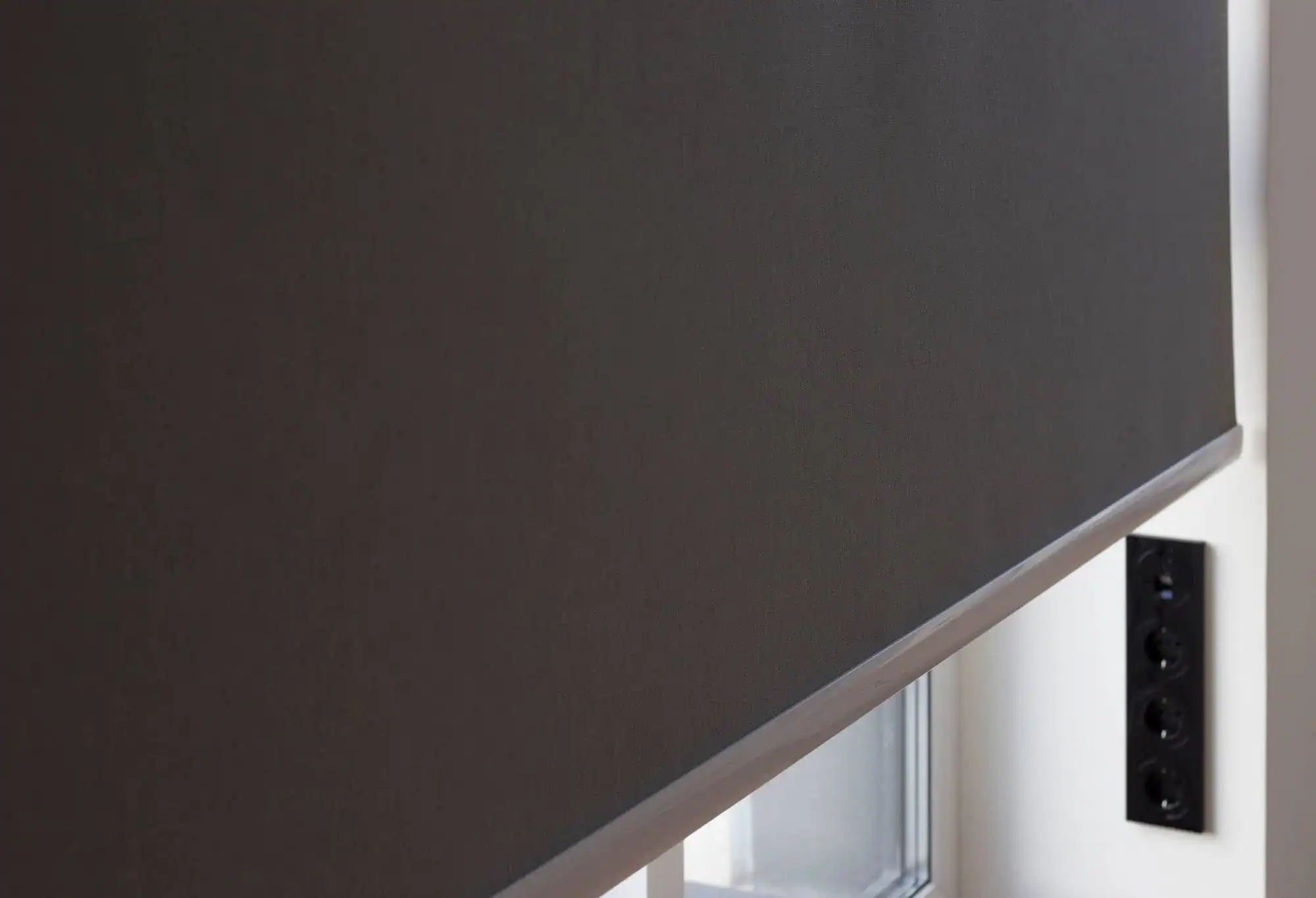 A close-up of a dark roller blind, a popular windows treatment in Miami-Dade County, FL, partially covering a window, with a white wall and a black panel featuring electrical outlets visible on the right side.