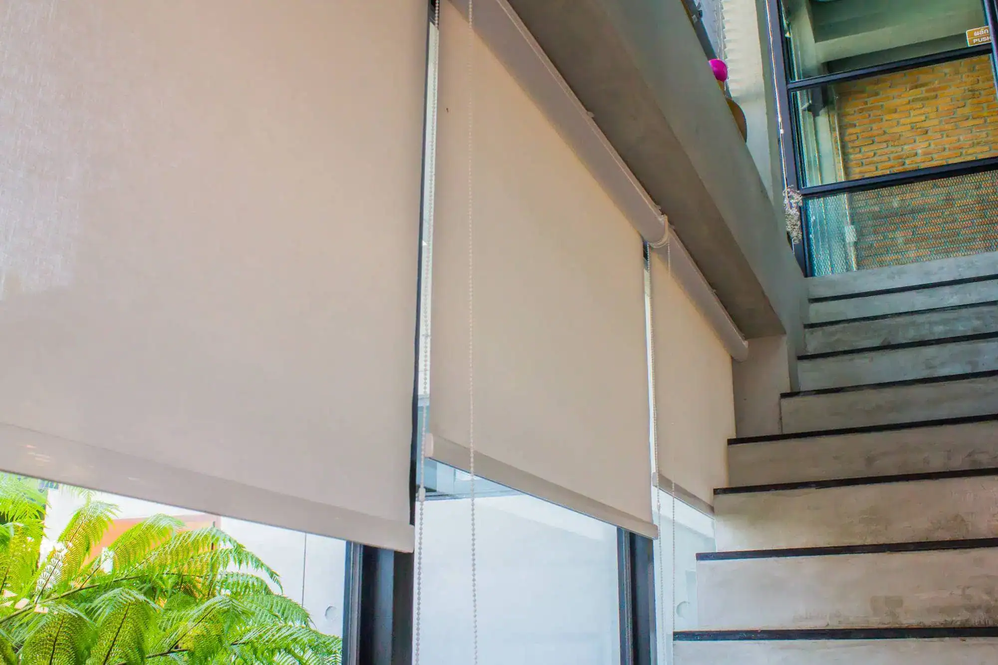 White roller blinds, a popular windows treatment in Miami-Dade County, are partially lowered over large glass windows beside a modern staircase with concrete steps; lush greenery can be seen outside.