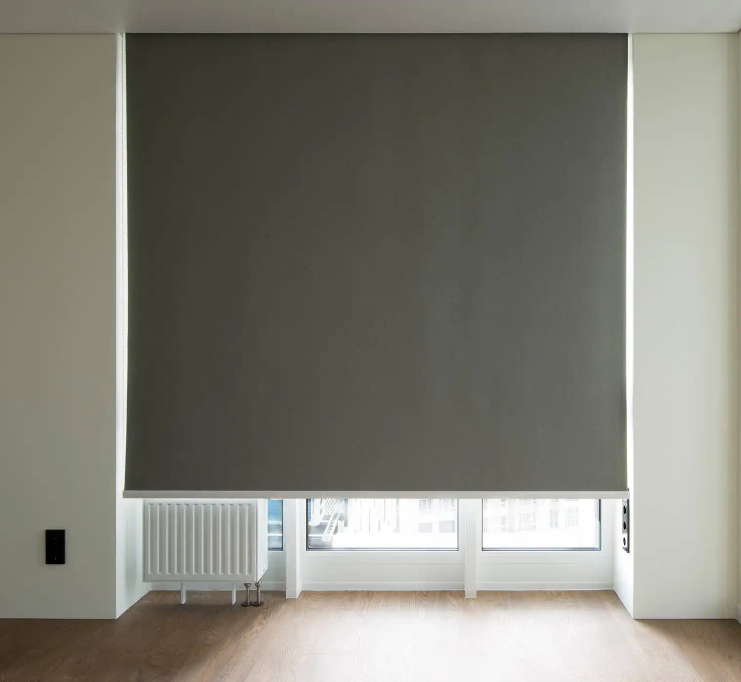 A large gray roller blind, a stylish example of windows treatments Miami-Dade County, FL residents love, is partially lowered over a window in a minimalist room with white walls, a radiator below the window, and light wood flooring.