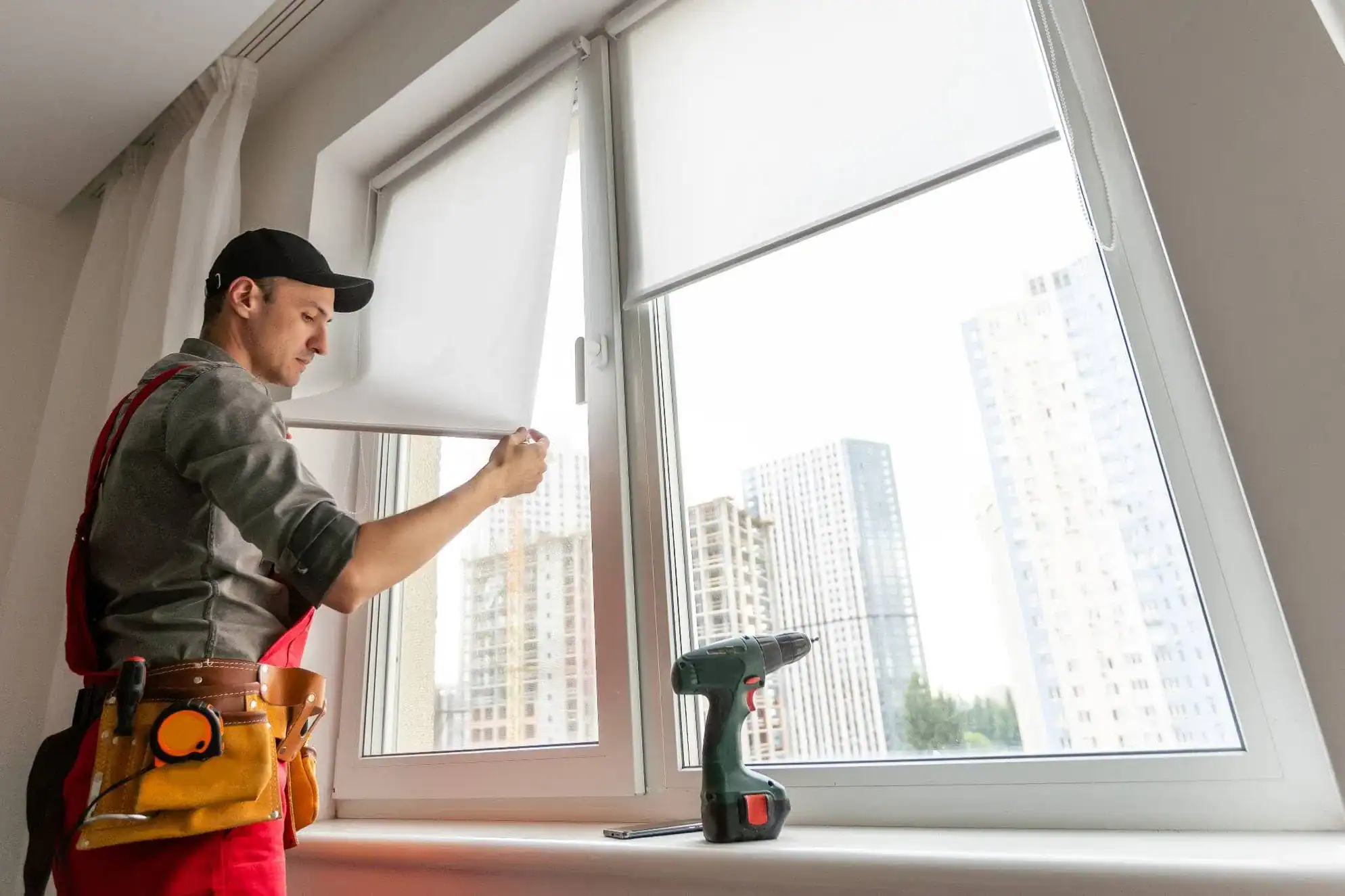 A worker in overalls and a cap adjusts a white roller blind on a large window, showcasing expert windows treatments Miami-Dade County, FL. A cordless drill rests on the sill, with tall buildings visible outside.