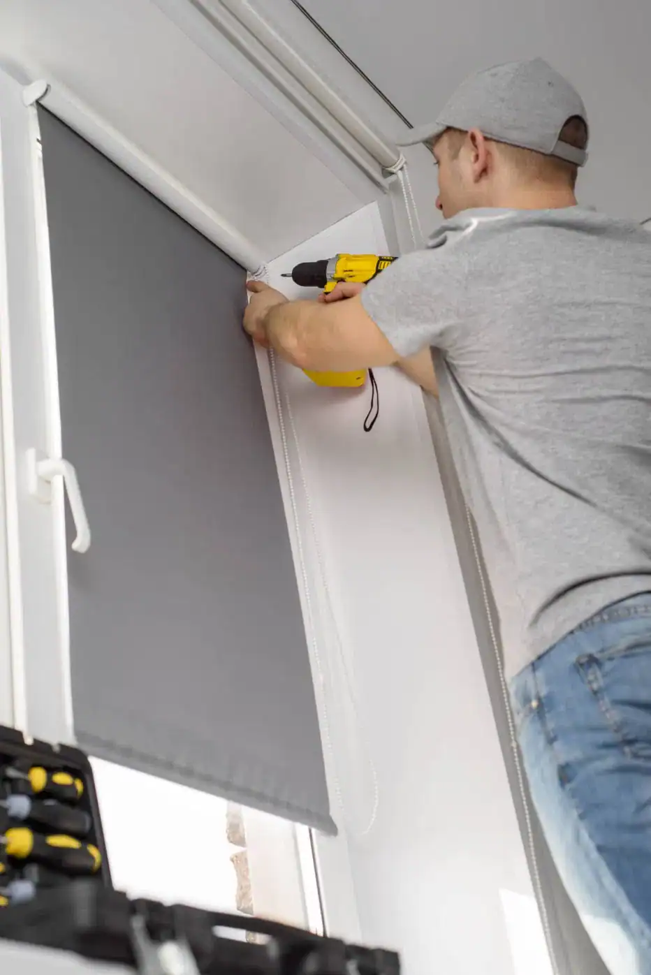A person in a gray shirt and cap uses a yellow power drill to install or repair window blinds—common windows treatments in Miami-Dade County, FL—inside a room. Tools are visible in a nearby toolbox.