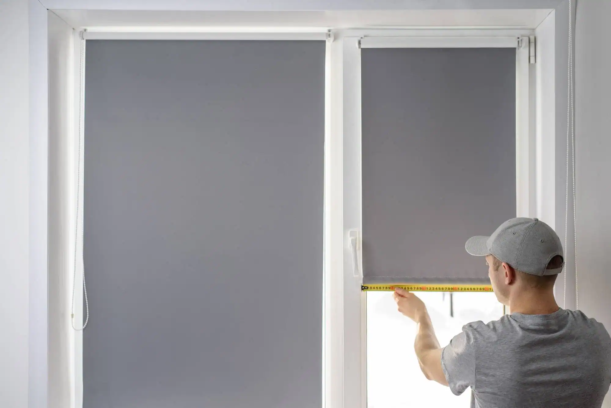 A person wearing a gray cap measures the width of a window covered with a gray roller blind using a yellow tape measure. The room, with white walls and another closed blind, reflects the precision of an FL home design company in Miami-Dade & Broward County.