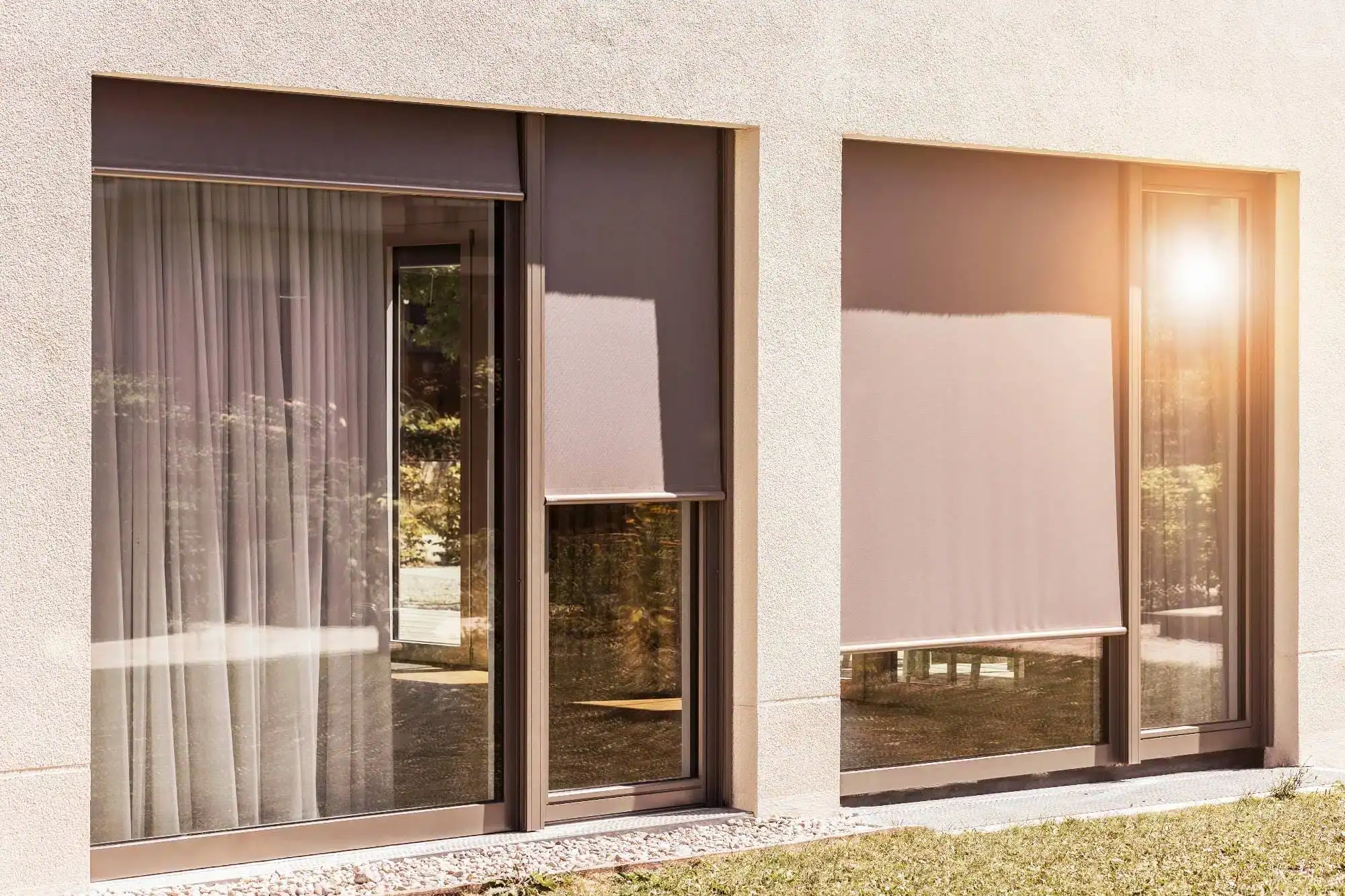 Two large windows on a beige building, designed by a home design company in Miami-Dade & Broward County, FL. One window has sheer curtains and the other a closed roller blind, sunlight reflecting off the glass with greenery visible outside.