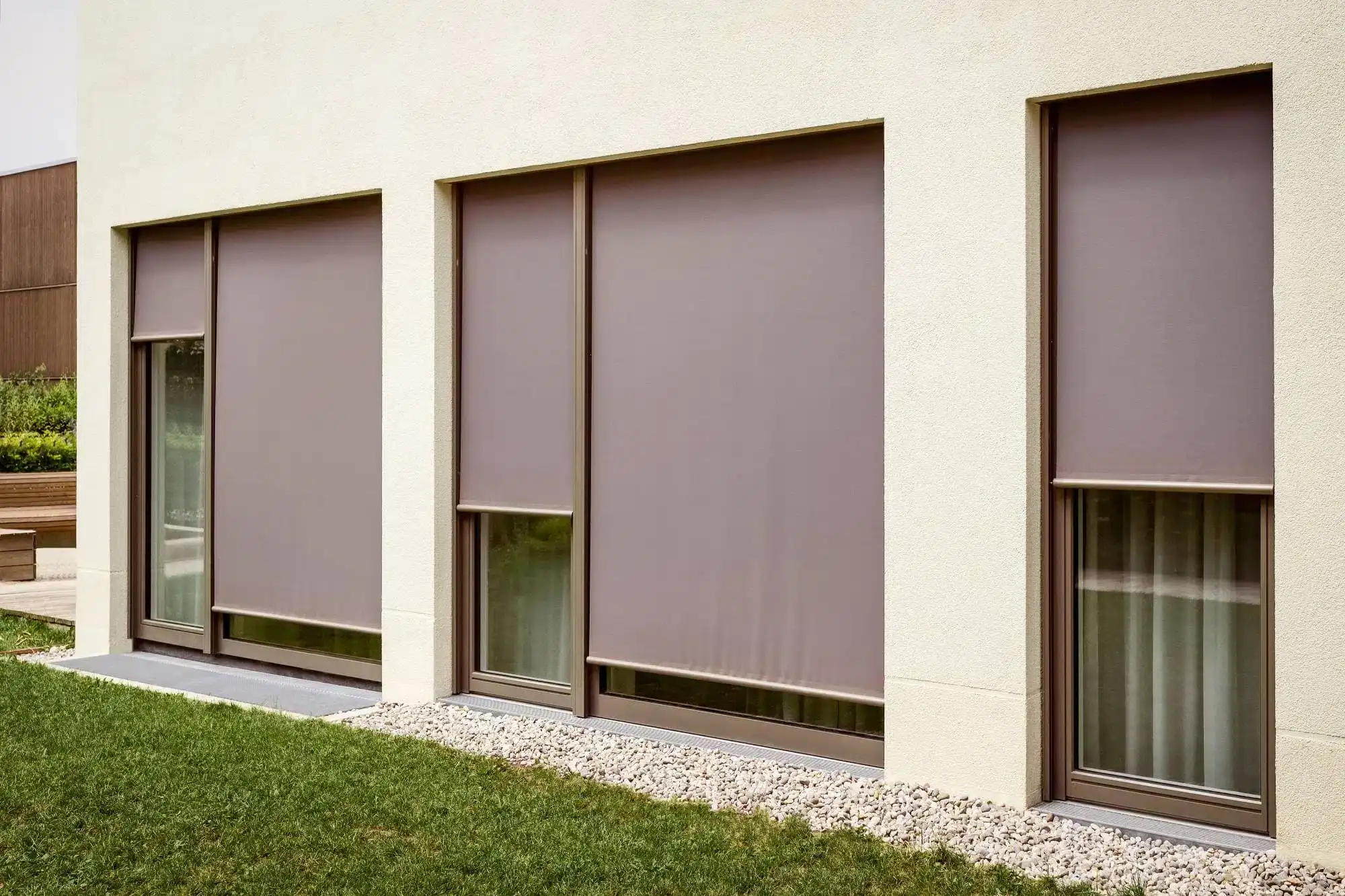 Modern house facade with three large rectangular windows, all covered by stylish brown exterior roller shades—an example of sleek windows treatments Miami-Dade County, FL loves. The windows sit above white gravel and a neatly trimmed green lawn.