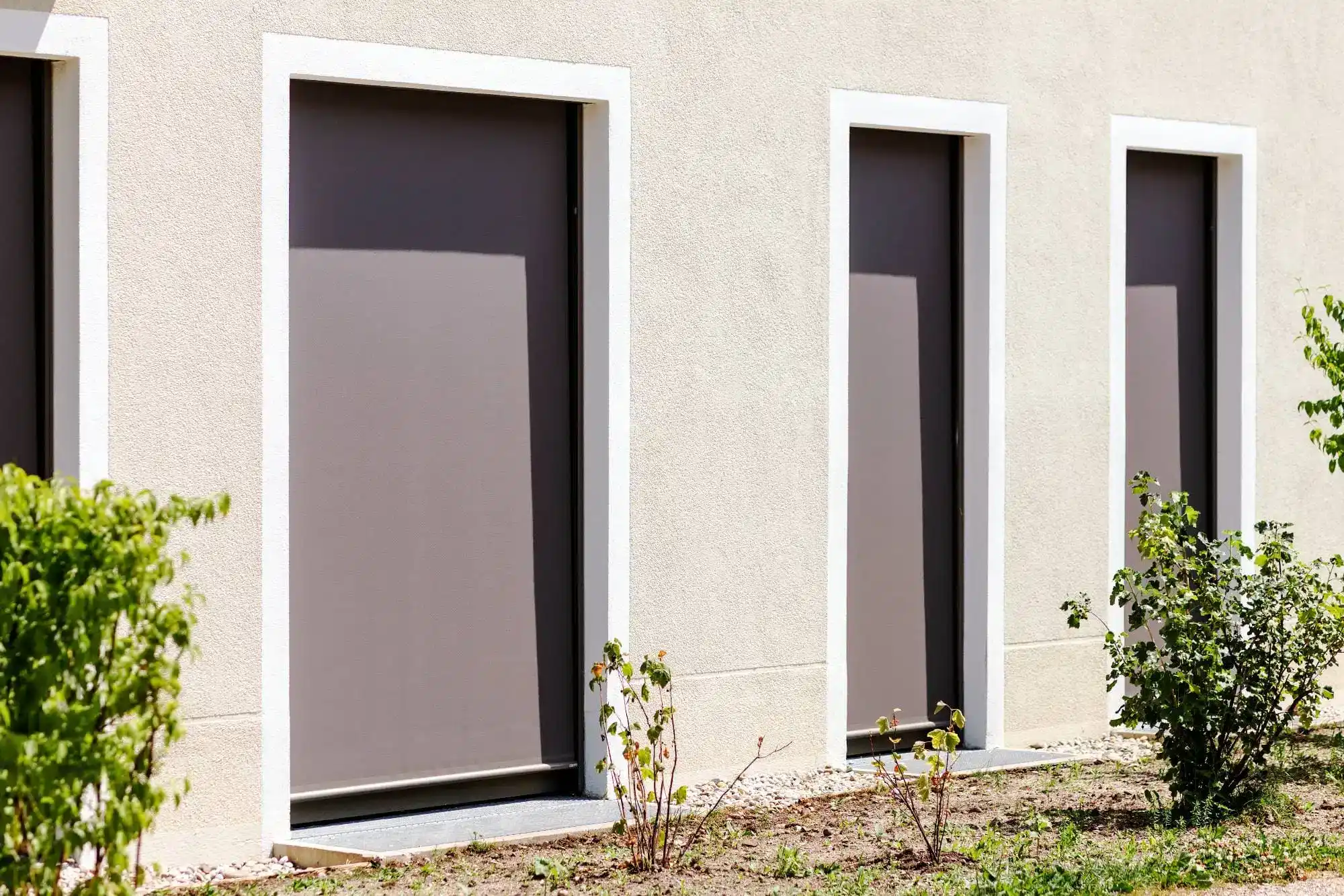 Three tall, rectangular windows with closed gray shutters—showcasing stylish windows treatments Miami-Dade County, FL—are set in a light-colored exterior wall. Small green bushes grow in the garden bed in front of the windows.