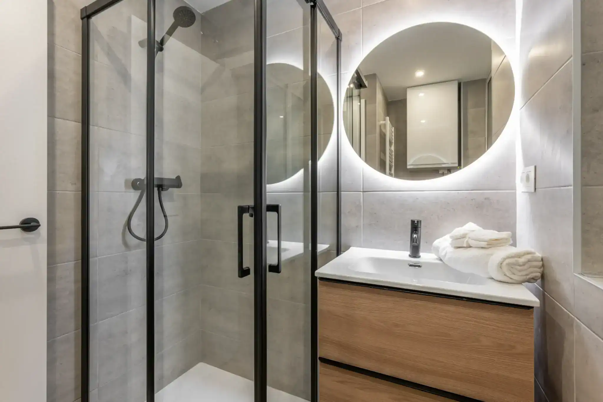 Modern bathroom with a glass shower enclosure, black fixtures, a wooden vanity and sink, a round illuminated mirror above the sink, neatly folded white towels on the countertop, and stylish windows treatments for Miami-Dade County, FL homes.