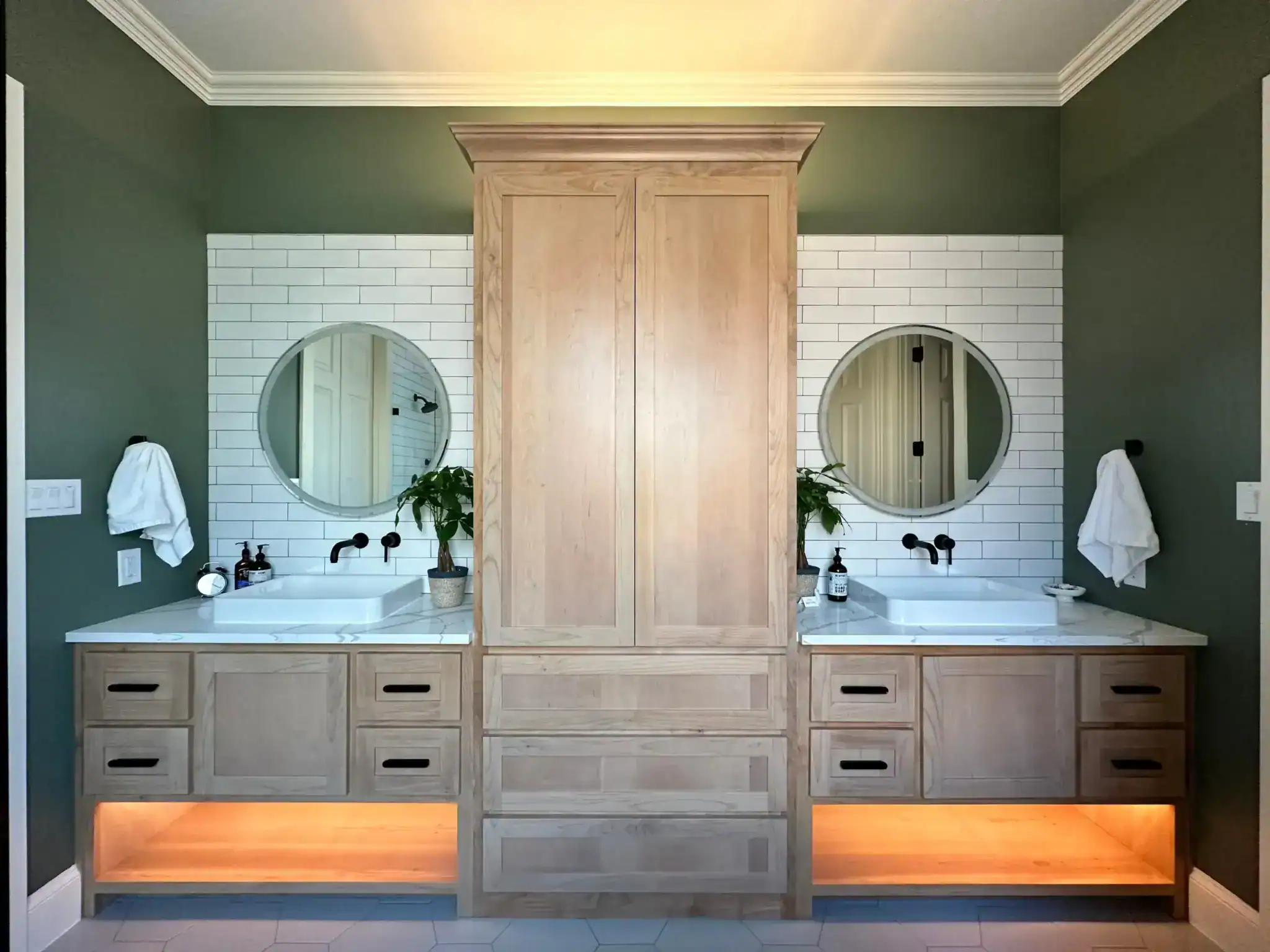 A modern bathroom with dual sinks, round mirrors, white subway tile backsplash, and a tall wooden cabinet in the center features stylish windows treatments Miami-Dade County, FL, plus plants, soap dispensers, towels, and warm under-cabinet lighting.