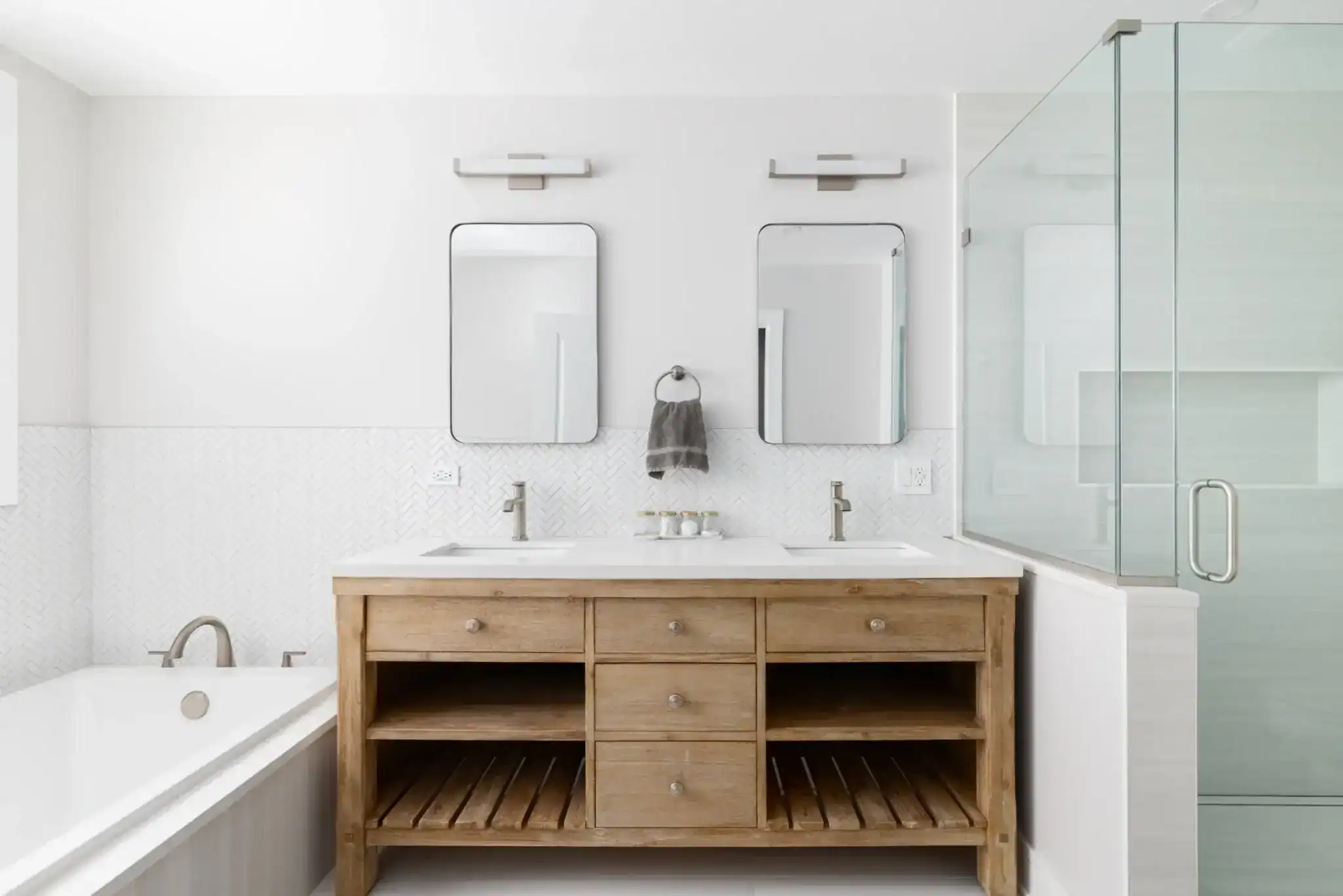 Modern bathroom remodeling in Miami Dade County FL featuring a wooden double-sink vanity, two rectangular mirrors, wall-mounted lights, a glass shower enclosure, and a bathtub on the left in a light, minimalist style.