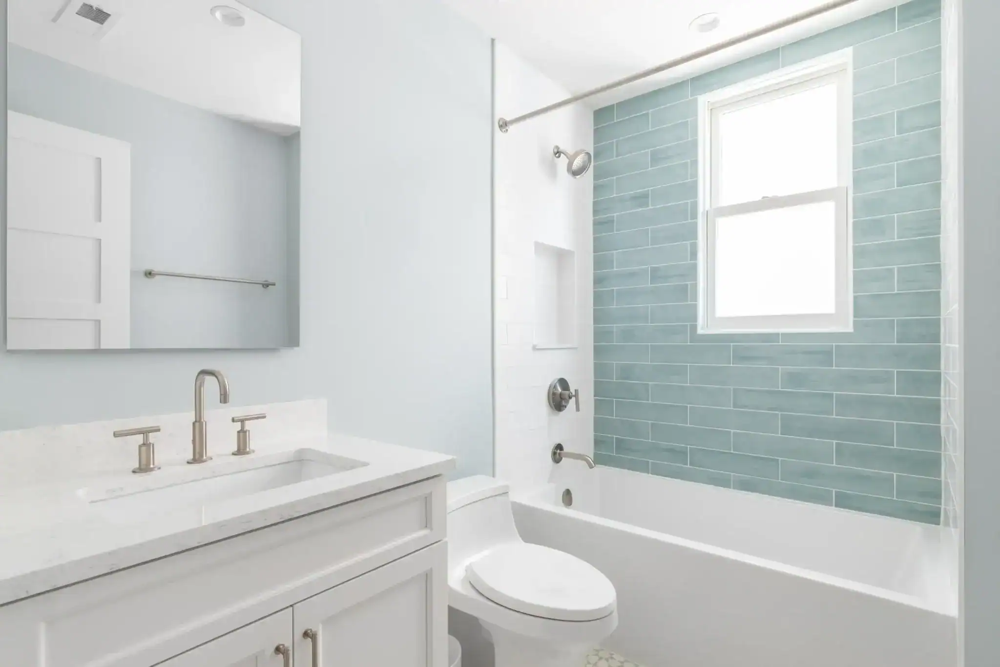 A bright, modern bathroom with a white vanity and sink, silver fixtures, a toilet, and a bathtub with blue subway tile beneath a large window featuring stylish windows treatments—perfect for any Miami-Dade County, FL home.