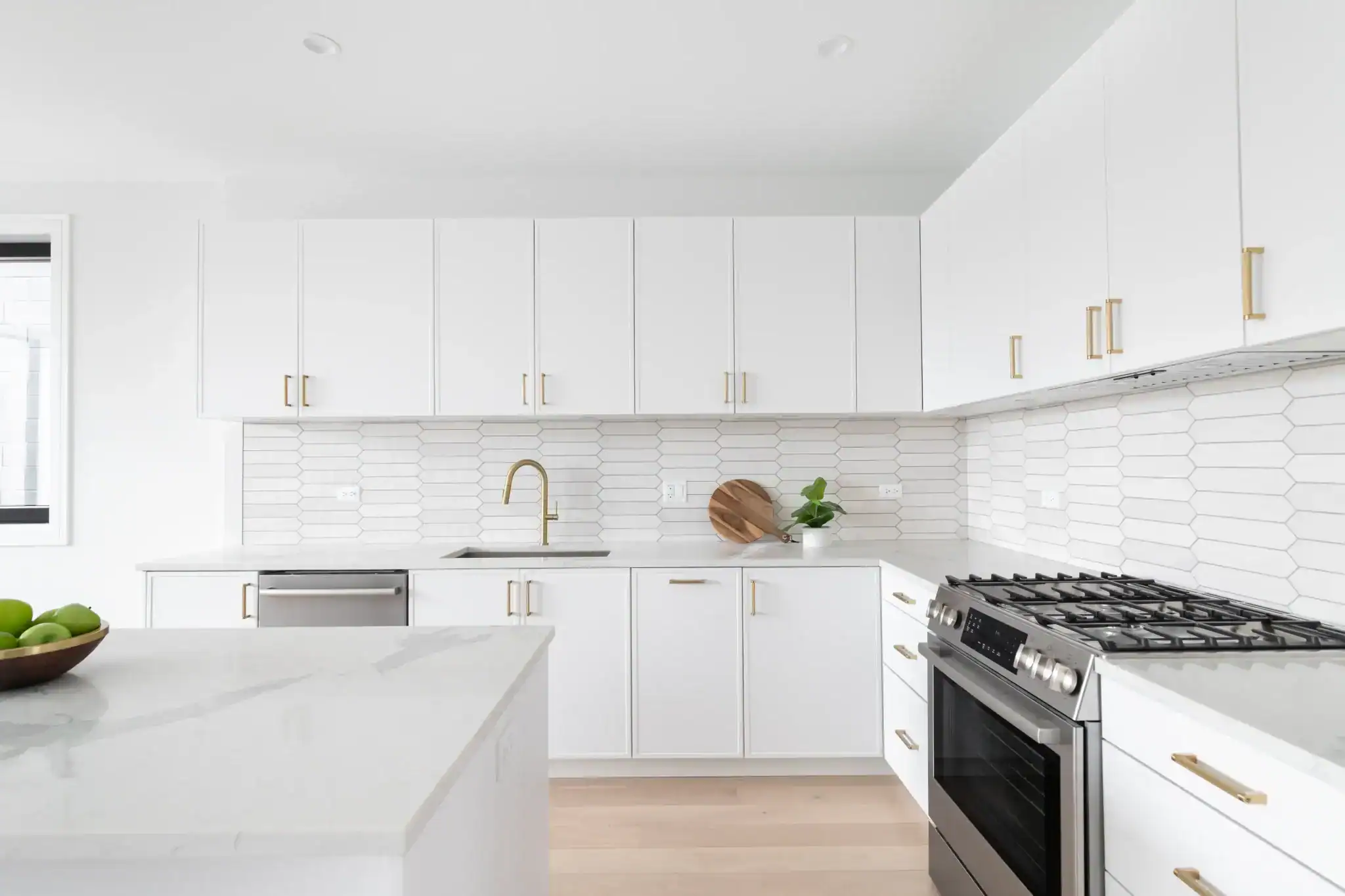 Modern FL kitchen with white cabinets, gold hardware, stainless steel appliances, and a sleek island featuring green apples. Elegant windows treatments Miami-Dade County complete the minimalistic decor and bright ambiance.