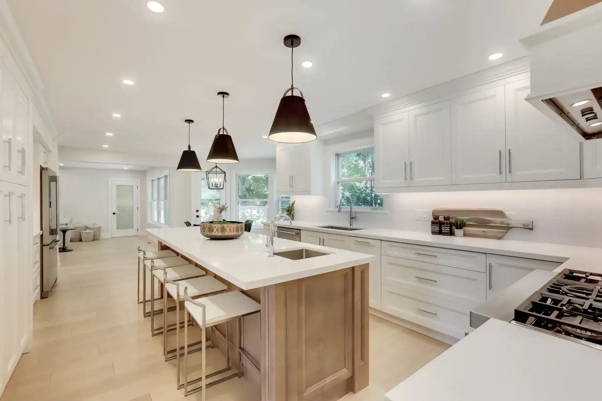 A modern kitchen designed by a top kitchen remodeler in Miami Dade County, FL, featuring white cabinets, a large island with a wooden base, four stools, pendant lights, stainless steel appliances, and expansive windows for natural light.