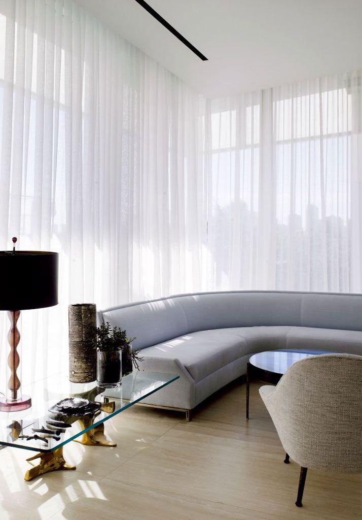 A modern living room with floor-to-ceiling sheer white curtains as windows treatments Miami-Dade County, FL, a curved light gray sofa, a glass coffee table with gold legs, a decorative lamp, and a light gray upholstered chair on a light wood floor.
