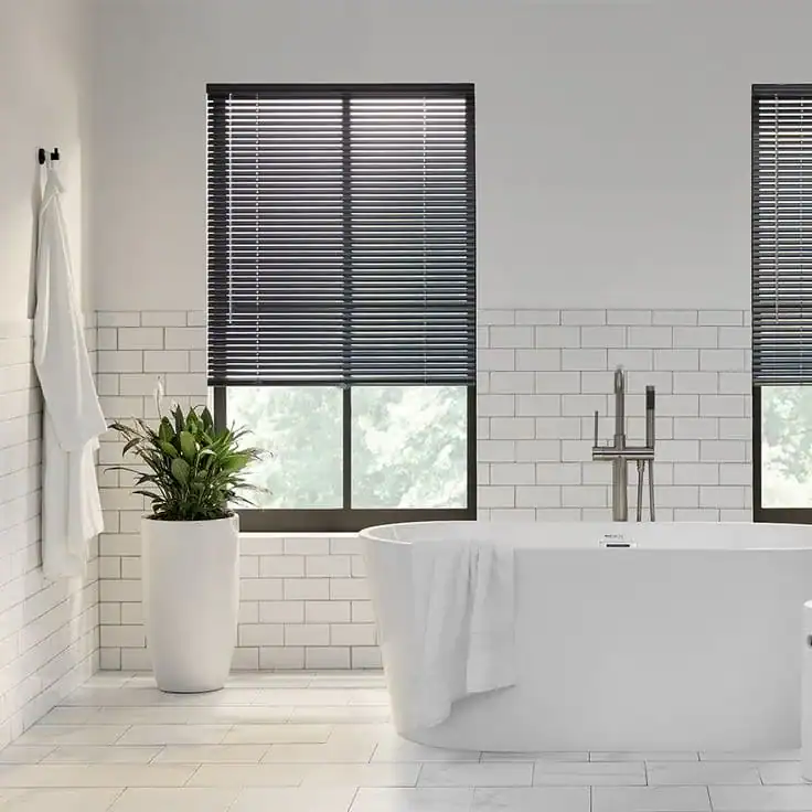 A modern bathroom with white tile walls and floor, a freestanding bathtub, a towel draped over the tub, a tall potted plant, and two large windows with dark blinds.