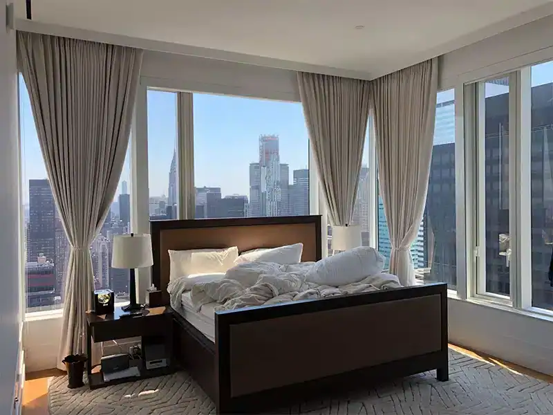 A modern bedroom with large windows offering a city skyline view, styled by a top home design company in Miami-Dade & Broward County, FL. Rumpled white bedding and light gray curtains complete the serene retreat, with a lamp on the bedside table atop a patterned rug.