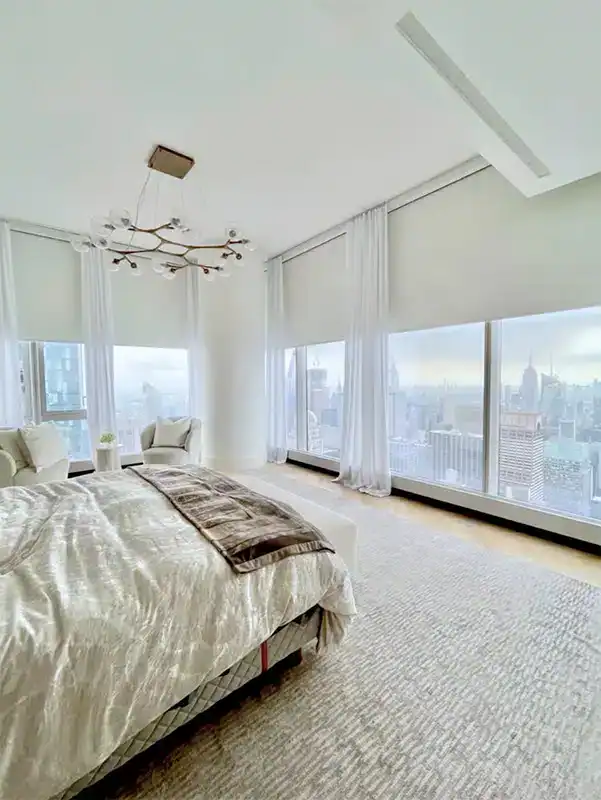 A modern, bright bedroom by a top home design company Miami-Dade & Broward County features floor-to-ceiling windows with city skyline views, a bed with light bedding, two armchairs, and an elegant chandelier on the ceiling.